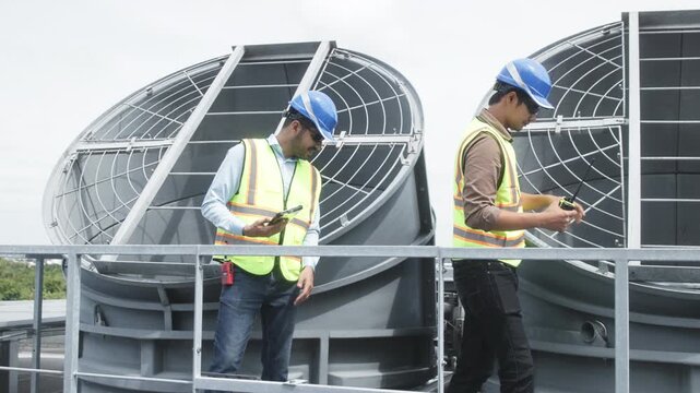 Two construction engineers in safety gear examine industrial HVAC units on rooftop, pointing and discussing project progress and setup.