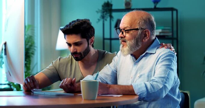 Indian young son teaching senior father how to use desktop computer at home in friendly way, elderly dad learning digital technology with smile while browsing internet together in modern living room