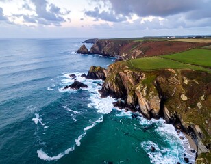 Aerial view of a rugged coastline meeting an ocean under a cloudy sky. Cliffs and foamy waves create a dramatic scene
