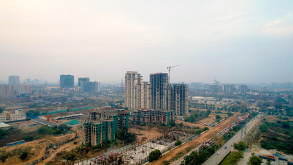 Naklejka premium drone view of high-rise residential buildings under construction with crane in Gurgaon with hazy skyline, highlighting urban growth, real estate development, and air pollution.