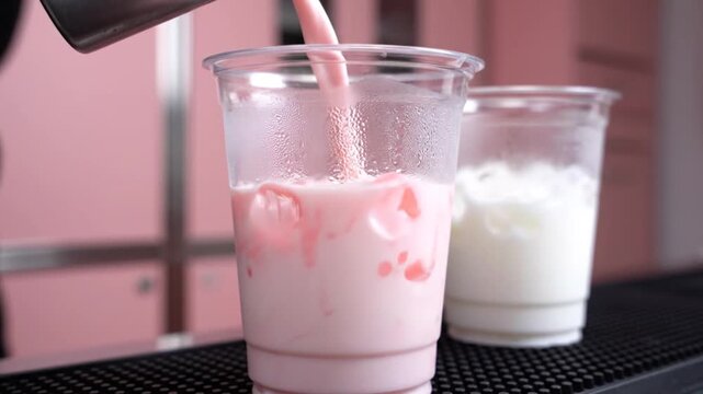 Pouring a pink beverage into a clear plastic cup, creating a creamy swirl with milk in another cup in the background