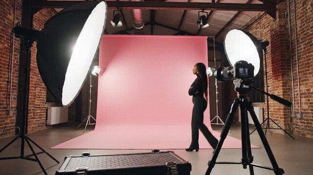 Woman in business attire walking in front of pink studio backdrop with professional photography equipment and softbox lights