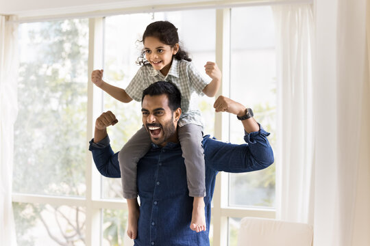 Athletic young Indian father carrying preschooler 6s adorable son child on his shoulders, screaming and showing strength of biceps muscles, perform strongman pose indoors. Sportive family, fatherhood