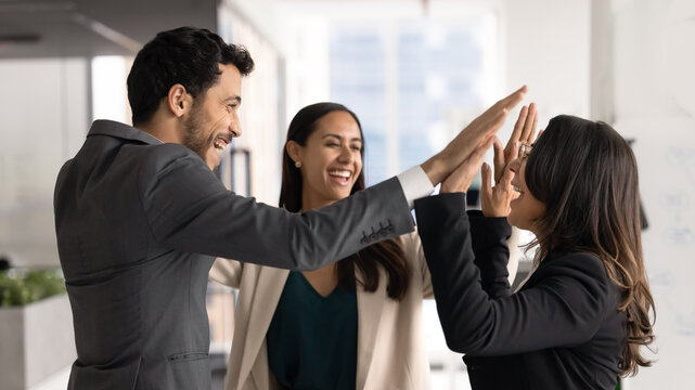 Three multinational happy teammates celebrate success, progress and career growth in office, exchanging high five gesture, enjoy project launch, task accomplishment. Triumph, achievement, team spirit
