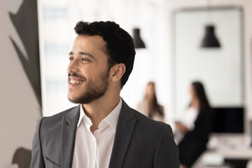Fototapeta premium Close-up of smiling Arabic man dressed in formal suit looking into distance with optimistic smile, dreaming about career perspectives, professional opportunity stands in modern workspace. Aspirations
