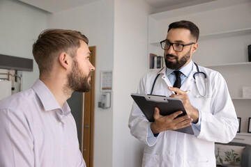 Collecting disease history. Attentive young male doctor in hospital uniform meet patient in office listen to complaints collect anamnesis fill in medical records by hand in paper form before checkup