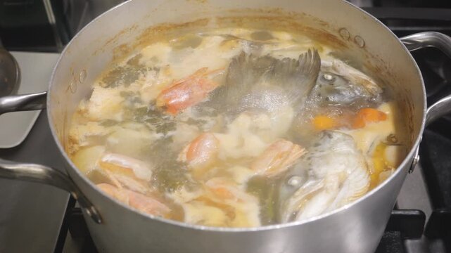 Seafood broth simmers in a pot with fish head, shrimp shell (Penaeus spp.), onion (Allium cepa), and herbs such as bay leaf (Laurus nobilis), creating a rich stock base, close up shot.