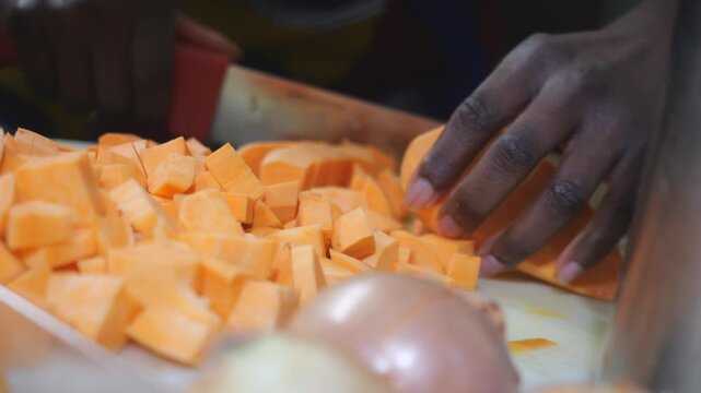 Diced yam cubes (Dioscorea spp.) pile up on a cutting board as an African woman chef chops more pieces with a large knife for velout&eacute; soup preparation in a kitchen, close up shot.