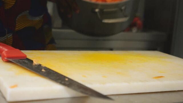 Close view of an African woman chef slicing peeled yam on a cutting board with a large kitchen knife, preparing cubes for a velout&eacute; style soup, with a stainless steel pot nearby in a working kitchen.