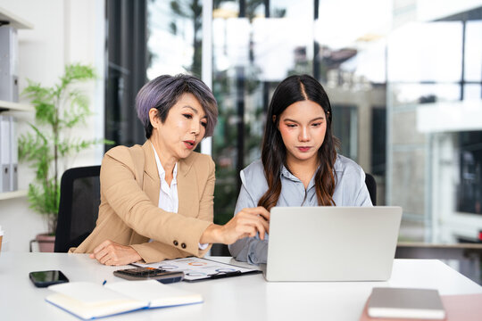 Confident mature business professional woman talking to younger female colleague at office table, speaking, gesturing, teaching, explaining work tasks. Elder mentor training intern.