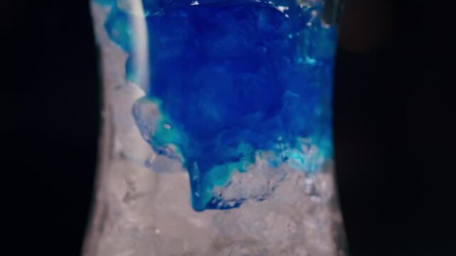 Alcoholic drink being poured over crystal-clear ice cubes in a cocktail glass against a dark background.