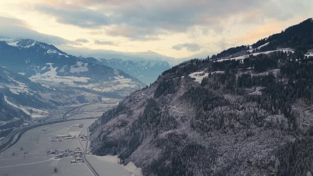 Aerial view of the Kaltenbach valley in Austria during sunrise, with soft light touching alpine meadows and mountain slopes. Morning mist lingers above fields while small houses and winding roads beco