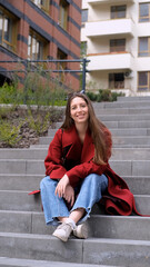 Portrait of a beautiful European woman in a red coat sitting on the steps of a city street