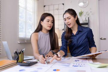 Obraz premium Two young Asian businesswomen reviewing financial reports and discussing data analysis in a bright modern office.