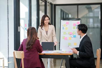 Fototapeta premium Group of young Asian professionals engaging in a dynamic brainstorming session. A female leader stands confidently while presenting ideas to her colleagues, showcasing a collaborative, positive