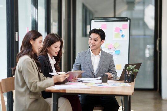 Asian business team, consisting of one man in a grey blazer and two women in blazers, are collaborating around a wooden table in a modern, sunlit office. They are happily discussing documents on a cli