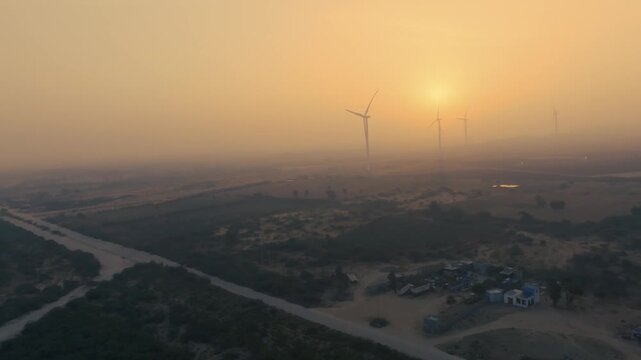 Cinematic aerial pull-back showing a wind turbine field and a dirt access road during a hazy morning. The blurred sun and thick fog create a powerful atmosphere.