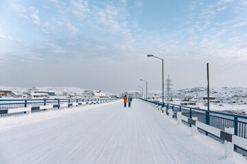 Obraz premium two girls in colored jackets are walking in winter on a snow covered empty bridge in a town in the far North