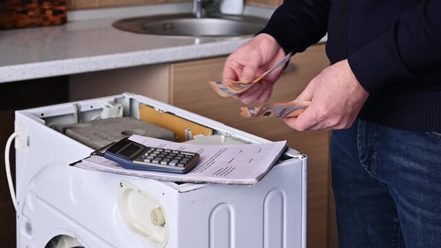 An adult man calculates the cost of a washing machine repair on a calculator and counts out euro bills. xpensive appliance repairs and maintenance.