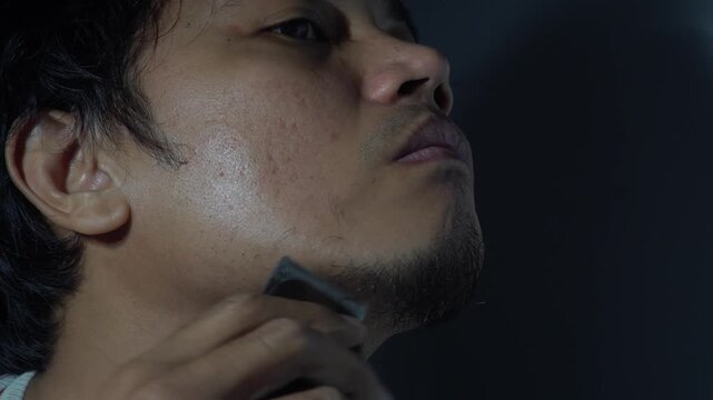 Side view close up of a young Asian man using an electric hair trimmer to groom his beard and chin. Detailed footage showing male facial skincare, morning grooming routine, and the use of modern elect