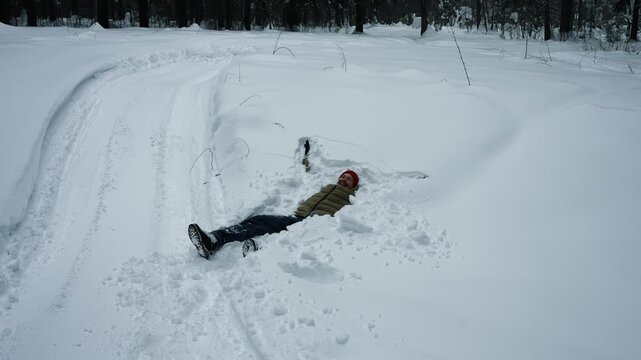 Cheerful young man wearing winter clothing having fun falling backward into a deep snowdrift and creating a perfect snow angel shape by waving his arms and legs in the fresh powder