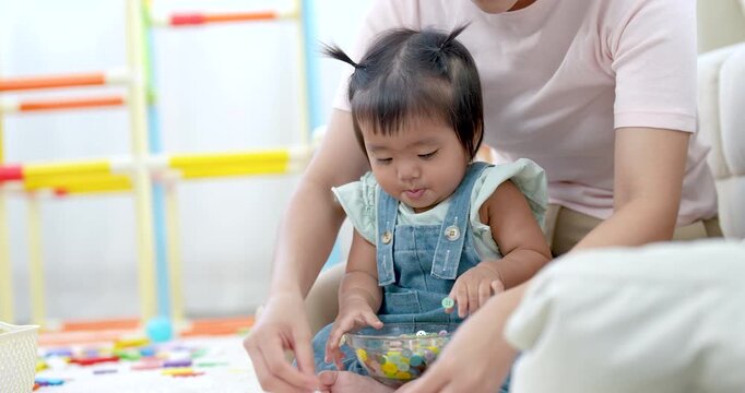 Asian toddler girl holding small beads in bowl while mother helps guide tiny hands, focused face, bright light across home, learning, bonding moment between parent and child, playful education