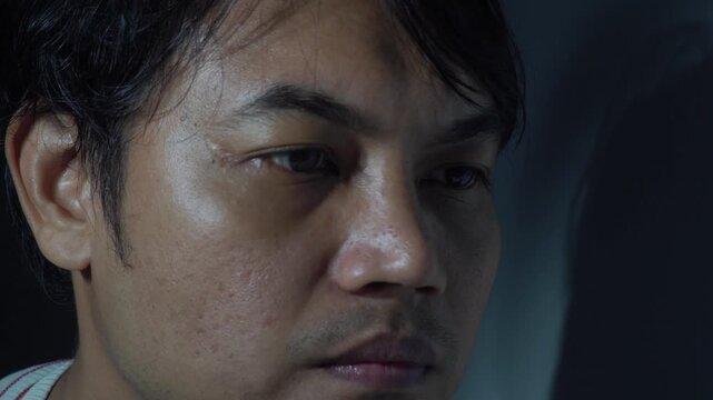 Close-up of a depressed Asian man with a sad and weary expression looking away in low light. Concept of mental health, anxiety, loneliness, sorrow, and psychological distress. Emotional cinematic port
