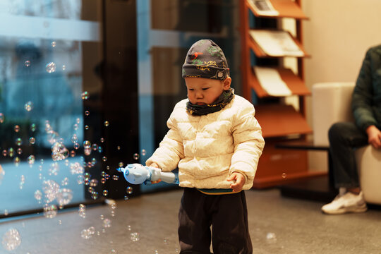 Asian boy playing with bubble machine indoors in winter wearing padded jacket and hat