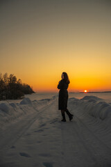 Obraz premium Young woman in dark dress stands on snow-covered landscape during sunset, with vibrant orange and yellow sky illuminating the scene behind her