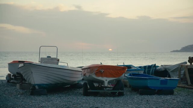 A row of small boats rests on a rocky beach as the sun sets over a calm sea, casting soft light across the coastal scene.