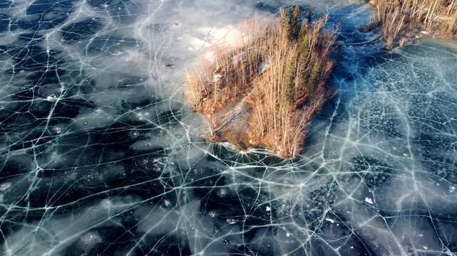 Aerial drone footage descending over Matanuska Lake, revealing a small rocky island dotted with scrubby dead trees and weathered branches. The frozen surface stretches endlessly beneath, with deep blu