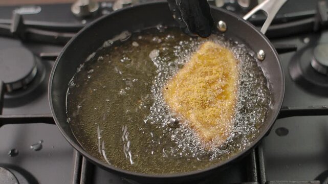 High angle close up of a perfectly breaded chicken cutlet sizzling in hot oil, lifted by tongs to reveal its crispy golden texture.
