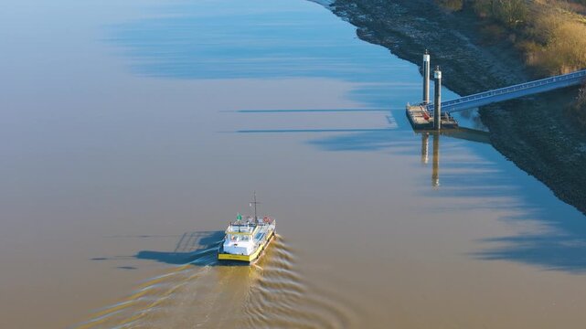 small yellow ferry cruising across calm scheldt river metal landing platform muddy banks tall pier pillars casting reflections sint amands belgium toward smooth drone view sint-amands 