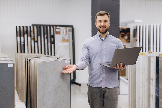 Salesman holding laptop and presenting flooring tiles, working in a modern showroom for interior design