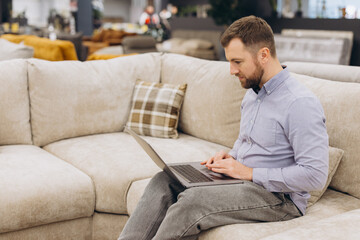 Fototapeta premium Man is seated on a contemporary couch, using a laptop in a showroom setting, exploring the ideas of remote work, home office setup, comfort, and technological integration
