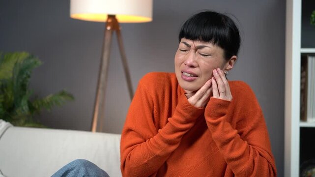Woman with toothache pain. Dental problem, sensitive teeth, healthcare and medicine concept. Asian female sitting on sofa, touching cheek and feeling discomfort. Dentistry and health care at home.