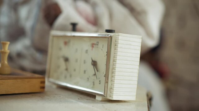 Close up of dark skinned dirty hand in white sleeve pressing mechanical chess clock during indoor match, showing dusty surface and worn timer, with wooden chessboard partially visible beside device