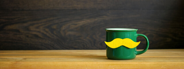 Green enamel mug with a yellow mustache decoration standing on a wooden surface, representing...