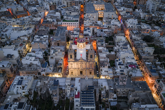 Aerial view of the illuminated Stella Maris Parish Church amidst the densely packed buildings, casting a warm glow against the cool evening hues, Sliema, Malta.