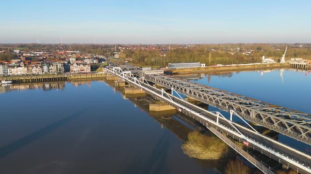 high altitude steel truss bridge stretching across calm reflective river connecting riverside town colorful buildings wooded banks under clear blue sky to long drone view temse belgium 