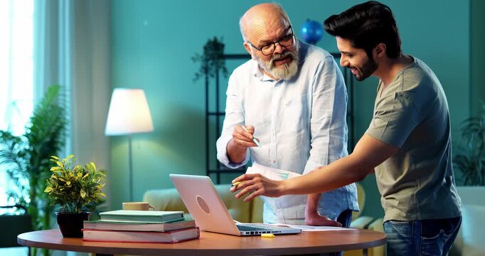 Indian young son and senior father filing online tax return together using laptop while standing at table in modern home, discussing financial reports, income statements yearly accounting documents