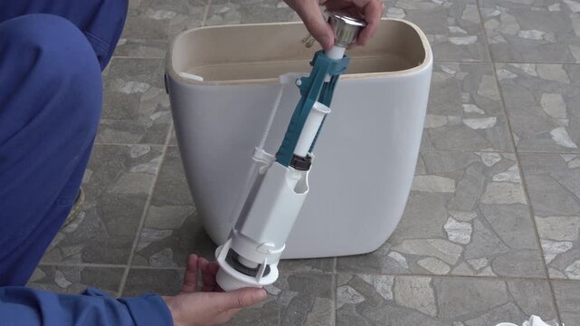 Plumber in blue uniform installing a flush valve mechanism into a ceramic toilet tank, Hands of a technician repairing a toilet cistern with new plumbing parts