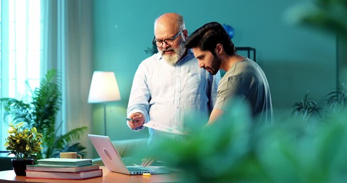 Indian young son and senior father filing online tax return together using laptop while standing at table in modern home, discussing financial reports, income statements yearly accounting documents
