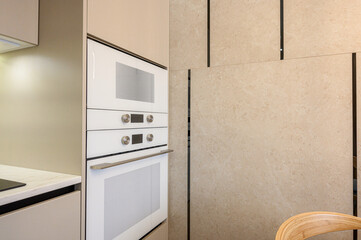 Fototapeta premium A modern kitchen with built-in white ovens, sleek metallic handles, beige cabinetry, marble-textured wall panels, and a minimalist design with clean lines.