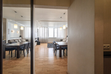 Fototapeta premium A modern living space with wooden flooring, two dining tables with dark chairs, pendant lights, sheer curtains, a sofa, and a cozy rug. Bright and airy atmosphere