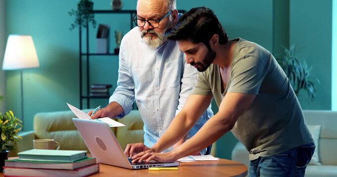 Indian young son and senior father filing online tax return together using laptop while standing at table in modern home, discussing financial reports, income statements yearly accounting documents