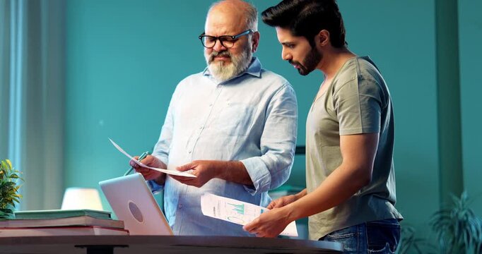 Indian young son and senior father filing online tax return together using laptop while standing at table in modern home, discussing financial reports, income statements yearly accounting documents