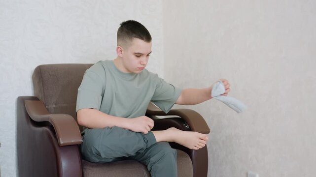 Kid sits on chair carefully pulling socks over feet, dressed in casual outfit, focused on getting ready indoors with neutral wall background and soft daylight creating calm morning routine scene