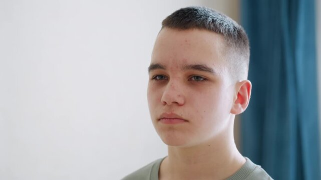 Close up of young boy with calm yet confident expression staring ahead in softly lit room, subtle acne and natural skin texture highlight authentic teenage look in quiet indoor setting