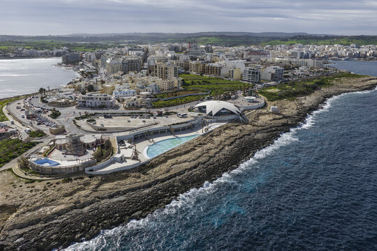 Aerial view of azure waters meet rugged coastline, framing the modern architecture and pools of St. Paul's Bay, Malta.